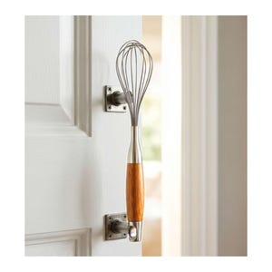 May include: A quirky door handle designed as a whisk. It has a stainless steel whisk head, a wooden grip, and stainless steel mounting hardware. The door is white, and the handle is mounted on a square plate.