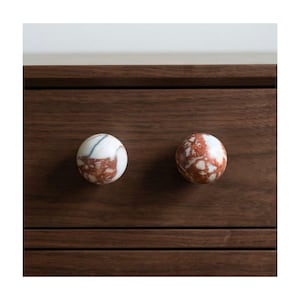 May include: Two marble cabinet knobs with a round shape. One knob is white with brown veining, and the other is red and brown. They are mounted on a dark brown wooden cabinet.