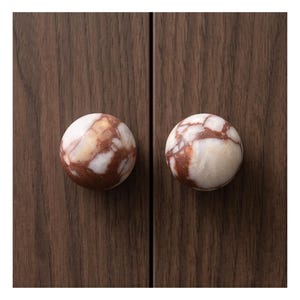 May include: Two round cabinet knobs with a marbleized appearance. The knobs are primarily white with brown veining and are mounted on a dark wood cabinet door. These decorative knobs add a touch of elegance to any furniture piece.