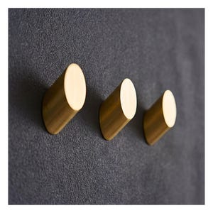 May include: Three gold-colored, cylindrical wall hooks mounted on a dark gray textured surface. The hooks are evenly spaced and angled slightly, offering a modern and minimalist design for hanging items. The hooks are made of metal.