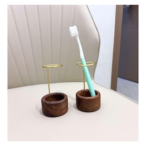 K&ouml;nnte beinhalten: Zwei Zahnb&uuml;rstenhalter. Jeder Halter hat einen dunkelbraunen Holzsockel und einen goldfarbenen Metallring. Ein Halter enth&auml;lt eine Zahnb&uuml;rste mit wei&szlig;em Kopf und hellgr&uuml;nem Griff. Die Halter stehen auf einer hellbeigen Oberfl&auml;che.