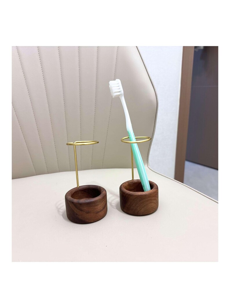 K&ouml;nnte beinhalten: Zwei Zahnb&uuml;rstenhalter. Jeder Halter hat einen dunkelbraunen Holzsockel und einen goldfarbenen Metallring. Ein Halter enth&auml;lt eine Zahnb&uuml;rste mit wei&szlig;em Kopf und hellgr&uuml;nem Griff. Die Halter stehen auf einer hellbeigen Oberfl&auml;che.