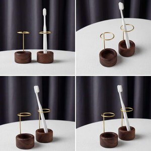 K&ouml;nnte beinhalten: Ein Set aus zwei Zahnb&uuml;rstenhaltern. Ein Halter ist ein kleiner, dunkelbrauner Holzzylinder mit einem goldfarbenen Metallring zur Aufnahme einer Zahnb&uuml;rste. Der andere ist ein passender Holzzylinder mit einem goldfarbenen Metallst&auml;nder.