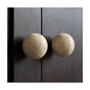 May include: Two round, beige cabinet knobs with a textured surface. The knobs are mounted on a dark brown wooden cabinet door. The knobs have a natural, stone-like appearance, adding a rustic touch to the cabinetry.
