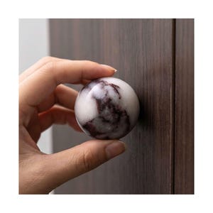 Könnte beinhalten: Ein Möbelknopf aus Marmor mit kugelförmiger Form. Der Knopf ist weiß mit dunkelburgunderfarbener Maserung. Er ist an einer dunkelbraunen Holzschranktür befestigt.