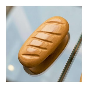 May include: A wooden bread-shaped door handle with diagonal grooves. The handle is light brown and appears to be made of solid wood. The handle is resting on a clear surface.