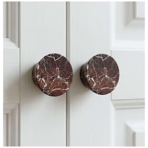 May include: Two circular cabinet knobs crafted from brown marble, featuring white veining. They are affixed to a white cabinet door. The knobs are cylindrical and have a polished finish, adding a touch of elegance to the cabinet.