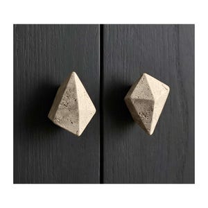 May include: Two diamond-shaped cabinet knobs made of a light beige stone-like material. The knobs are mounted on a dark grey wooden cabinet door. The knobs have a geometric, faceted design.