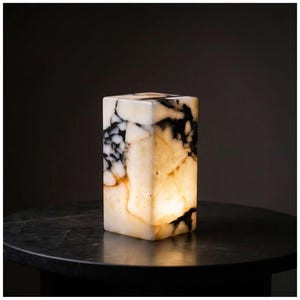 May include: A square onyx lamp, lit from within, displaying a striking pattern of black and white veining. The lamp emits a warm glow. It is positioned on a dark, round table, creating a contrast of light and shadow.