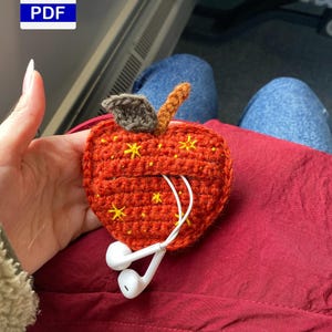 Op de afbeelding: Een oranje gehaakte appelvormige hoes met gele sterren, met witte oortelefoons. De appel heeft een bruin blad en een oranje steel. Het PDF-pictogram staat linksboven. Handgemaakt.