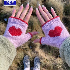May include: Pink knitted fingerless gloves with a red heart motif. The gloves have a red trim and are displayed against a backdrop of long grass. A PDF icon is also visible in the image.