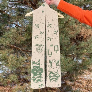 May include: A beige knitted scarf with green geometric and floral patterns. The scarf is hanging on a wooden hanger. The design includes stylized flowers, shapes, and a heart. The scarf is displayed against a backdrop of evergreen trees.
