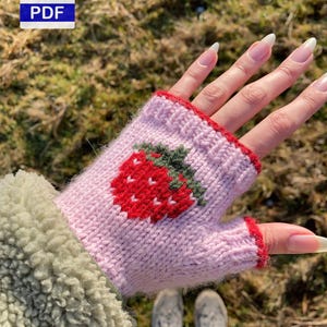 Könnte beinhalten: Rosa gestrickter fingerloser Handschuh mit einem roten Erdbeerdesign. Die Erdbeere hat einen grünen Stiel und Blätter. Der Handschuh hat eine rote Einfassung und wird an einer Hand getragen.