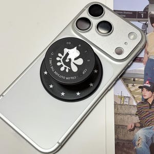 Puede incluir: Un teléfono inteligente plateado con un sistema de cámara de tres lentes y un agarre circular negro para teléfono adherido a la parte posterior. El agarre presenta un gráfico abstracto blanco, el texto "COLOR OUTSIDE THE LINES" y un borde de estrellas blancas.