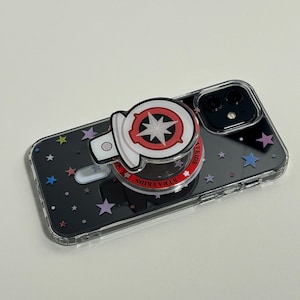 May include: A clear phone case with a black background and colourful star patterns. A white and red pop socket with a star design is attached. The phone is a black iPhone.