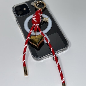 Puede incluir: Una funda transparente para teléfono con un cordón a rayas rojas y blancas. El cordón tiene detalles metálicos dorados y un dije de corazón dorado con la palabra "Stay" grabada. También hay un dije de estrella dorada.