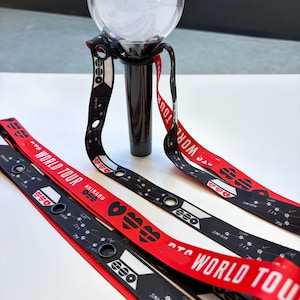Op de afbeelding: Een zwarte en rode lichtstick met een heldere bol aan de bovenkant. De lichtstick heeft een zwarte handgreep en rode en zwarte bandjes met de woorden "WORLD TOUR" en andere tekst. De bandjes hebben een hartvorm.