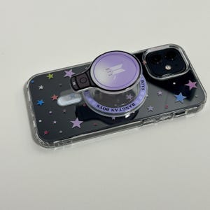 May include: A clear phone case with a black background and colorful star shapes. The case has a purple pop-up grip with the text "BTS" and a logo. The grip also has the words "BANGTAN BOYS" around the edge.