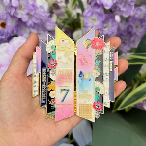 May include: A multi-coloured enamel pin with a stylized design, floral accents, and musical notes. The pin features the number 7 and the words "Forever" and "Butter". The pin is held in a hand against a backdrop of purple flowers.