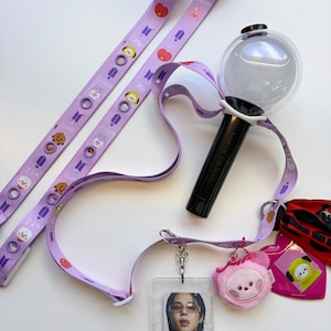May include: A collection of purple lanyards, a black and clear light stick, and a clear ID holder with a photo. The lanyards feature cartoon characters and the text "BTS". A pink plush charm and a pink tag are also included.