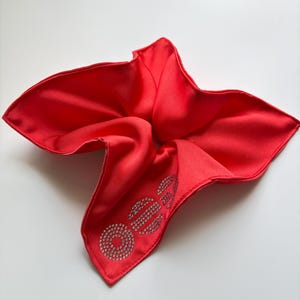 Pode incluir: Um lenço de cetim vermelho vibrante, com um design de strass brilhante. O design soletra a palavra "OEE" em uma fonte estilizada. O lenço tem formato quadrado.