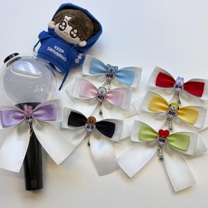 May include: A selection of satin bows in various colours, each with a character charm, and a plush doll wearing a blue hoodie with the text "KEEP SWIMMING". Bow colours include blue, pink, red, yellow, green, black, and purple.