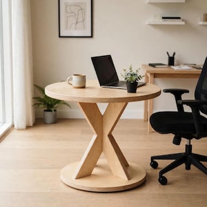 Puede incluir: Una mesa redonda de madera clara con una base en forma de X y un pie circular. La superficie de la mesa sostiene un portátil negro, una pequeña planta verde en maceta y una taza de cerámica beige. Una silla de oficina negra está junto a la mesa.