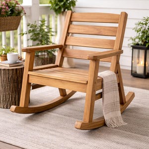 Puede incluir: Una mecedora de madera de color marrón claro con respaldo de listones y reposabrazos. Una manta doblada descansa sobre la silla. La silla está sobre una alfombra de color neutro. La escena está ambientada en un porche con plantas y una mesa pequeña.