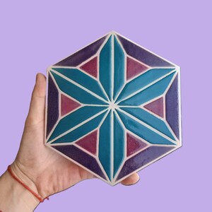 Op de afbeelding: Een zeshoekige tegel met een geometrisch sterrenpatroon. De tegel heeft een centrale turquoise ster omgeven door paarse en magenta driehoeken, allemaal omlijnd in wit. De tegel wordt tegen een lavendelkleurige achtergrond gehouden.