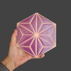Könnte beinhalten: Eine sechseckige Fliese mit einem geometrischen Sternmuster. Die Fliese hat eine glänzende lila Farbe mit hellbeigen Akzenten. Die Fliese wird in einer Hand vor einem dunkelgrauen Hintergrund gehalten.