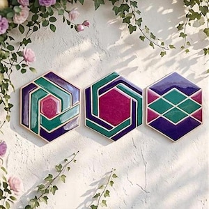 Può includere: Tre sottobicchieri esagonali con disegni geometrici. Ogni sottobicchiere presenta un motivo diverso nelle tonalità del viola, del verde acqua e del magenta, con un bordo color oro. I sottobicchieri sono esposti su una parete bianca strutturata con accenti floreali.