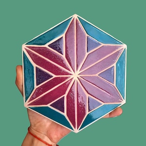 May include: A hexagonal tile with a starburst pattern. The tile has a teal border and a central star design in shades of purple and magenta. The symmetrical design is held in a hand.