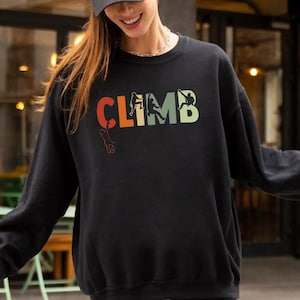 May include: Black crew neck sweatshirt with the word "CLIMB" in colorful letters. The design includes a climber silhouette and climbing gear. The sweatshirt is worn by a person outdoors.