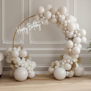 May include: A gold metal arch decorated with white balloons and floral accents. The words "Happy Birthday" are written in a cursive font on the arch. The arch is set up in a room with a couch, plant, and wooden table.