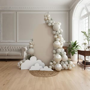 May include: A decorative backdrop featuring a large, neutral-toned oval panel adorned with a balloon garland in shades of white and cream. White cloud cutouts and a woven mat are at the base, creating a whimsical setting. The scene is set in a room with a sofa and large windows.