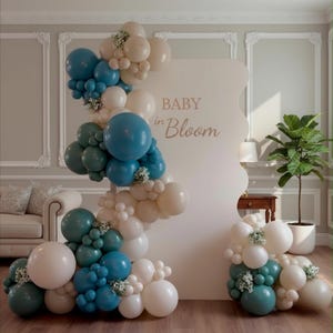 An elegant 'Baby in Bloom' balloon garland for a baby shower, featuring dusty blue, eucalyptus green, sand, and white balloons in various sizes framing a decorative welcome sign in a chic room.