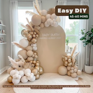 May include: A beige arch backdrop with the text "BABY SMITH ARRIVING AUGUST 2025" is decorated with a balloon garland in white, beige, and gold. The words "Easy DIY" and "45-60 MINS" are also visible. The setup is on a white rug.