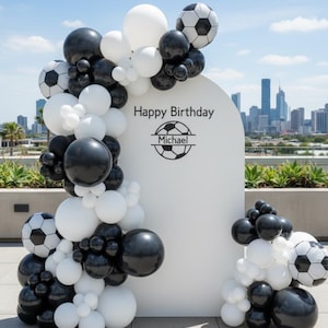 May include: A white arch backdrop with "Happy Birthday" text and a soccer ball graphic, decorated with black, white, and soccer ball-themed balloons. The balloons are arranged in clusters and arches, creating a festive atmosphere.