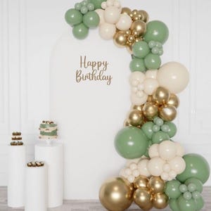 May include: A birthday party scene with a white backdrop and a balloon arch in shades of green, gold, and cream. The words "Happy Birthday" are written on the backdrop. White pedestals hold a small cake and cupcakes.