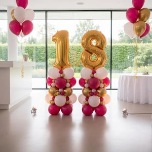 May include: Two balloon columns featuring large gold foil number balloons forming the number 18. Each column is constructed from a base of smaller white, magenta, and gold balloons. Additional balloon clusters in white, magenta, and gold float.