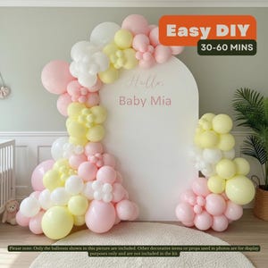 May include: A pastel balloon arch kit with pink, yellow, and white balloons. The arch frames a white backdrop with the text "Hello Baby Mia." The image also includes the text "Easy DIY" and "30-60 MINS."