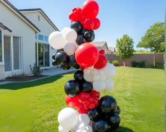 May include: A vertical balloon garland in red, black, and white, arranged in a cascading design. The balloons vary in size, creating a dynamic and festive display. The garland is set outdoors on a green lawn.