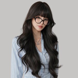 May include: A person wearing black-framed glasses and a light blue striped shirt. The person has long, dark wavy hair with bangs and a silver necklace. The background is a solid light gray.