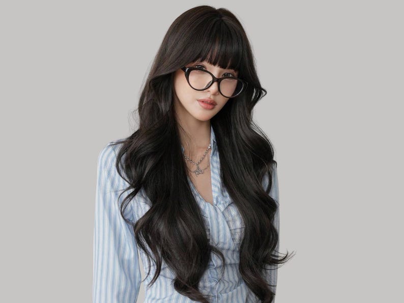 May include: A person wearing black-framed glasses and a light blue striped shirt. The person has long, dark wavy hair with bangs and a silver necklace. The background is a solid light gray.
