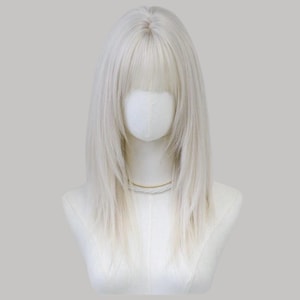 Puede incluir: Una peluca larga, rubia platino, con flequillo recto, exhibida sobre una cabeza y hombros de maniquí blancos. La peluca tiene una línea de cabello de aspecto natural y un brillo sutil. Se ve un delicado collar dorado.