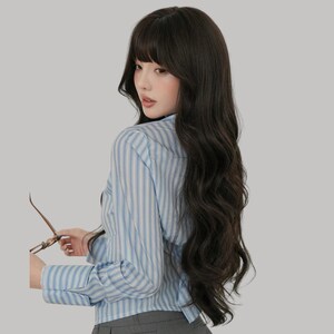 May include: A woman with long, dark wavy hair and bangs, wearing a light blue and white striped button-down shirt. She is holding a pair of eyeglasses. The background is a solid light gray.