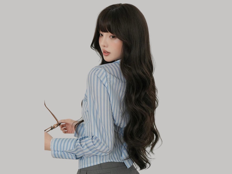 May include: A woman with long, dark wavy hair and bangs, wearing a light blue and white striped button-down shirt. She is holding a pair of eyeglasses. The background is a solid light gray.