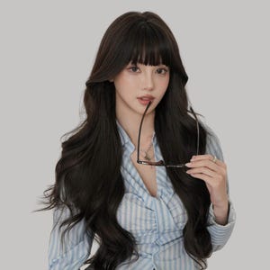 May include: A person with long, dark hair and bangs, wearing a light blue striped shirt, holding eyeglasses. The background is a solid, neutral gray.