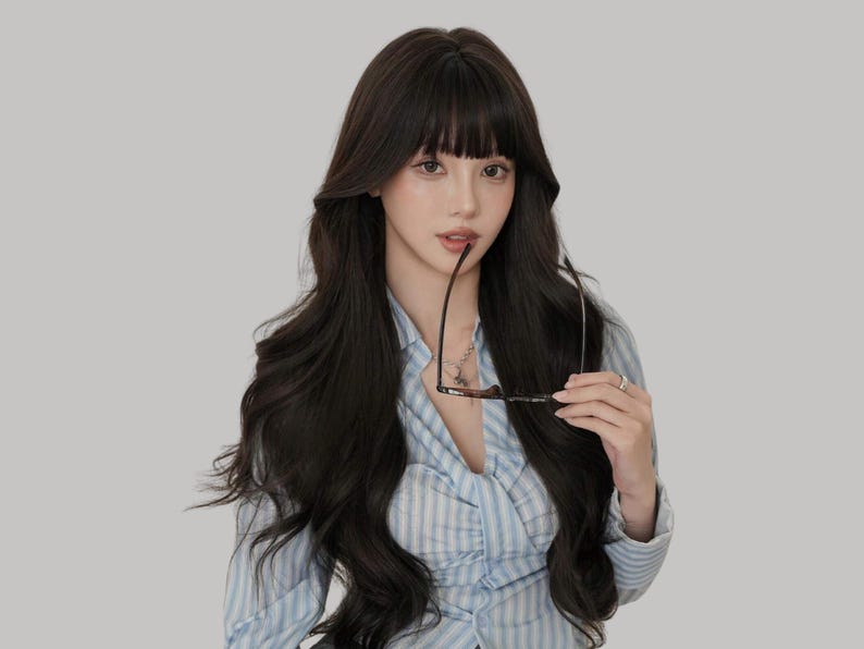 May include: A person with long, dark hair and bangs, wearing a light blue striped shirt, holding eyeglasses. The background is a solid, neutral gray.