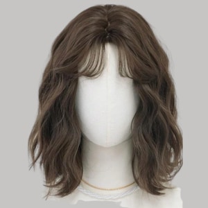May include: A brown wig with wavy, shoulder-length hair and bangs. The wig is styled on a mannequin head with a neutral background. The hair has a natural look and texture, suitable for everyday wear or cosplay.